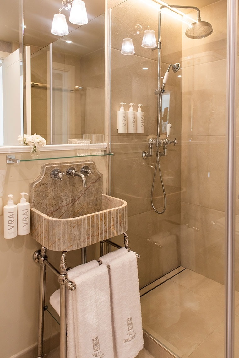 Banheiro de hotel Villa Dorotea com pia de mármore, ducha, toalhas e amenidades de luxo VRAI.
