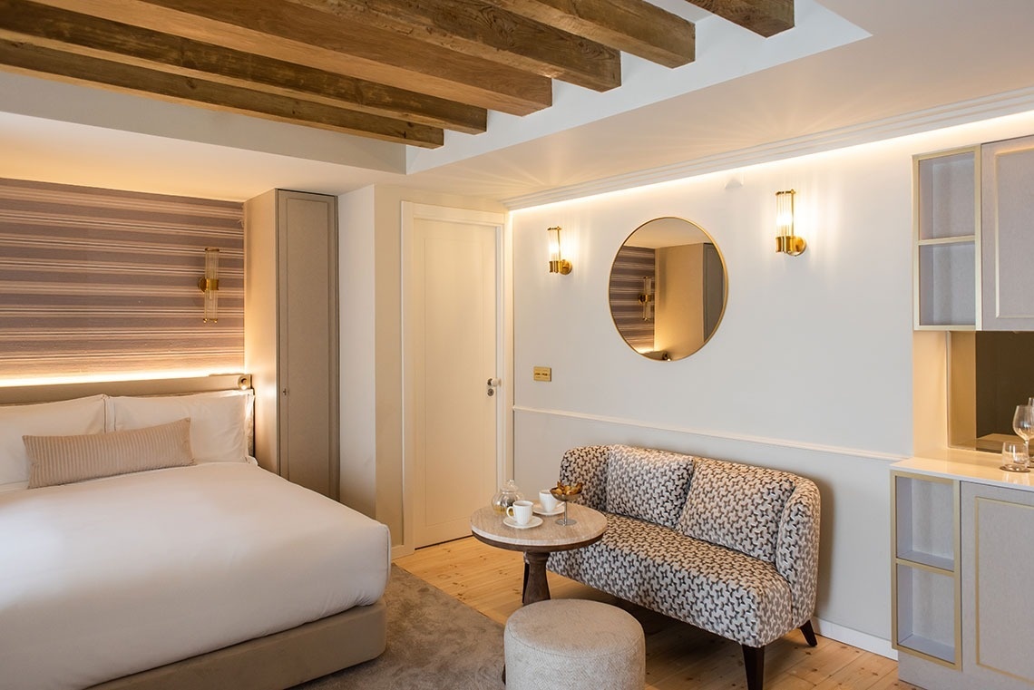 Aconchegante quarto de hotel com cama confortável, sofá, teto de vigas de madeira e iluminação ambiente.
