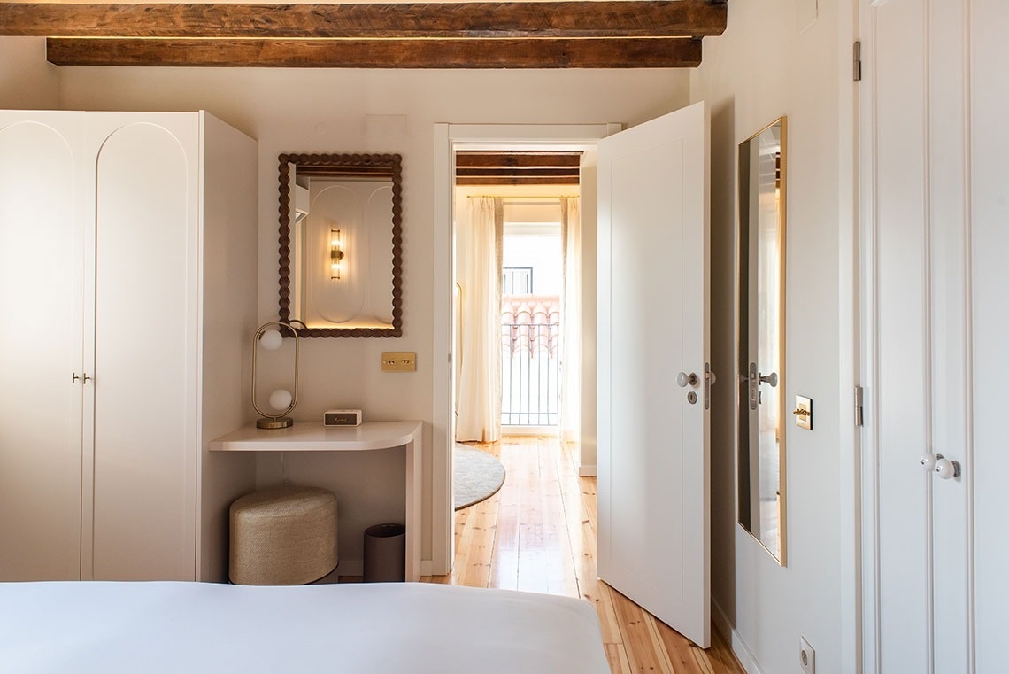 Quarto de hotel moderno e luminoso com varanda, toucador, armário branco e detalhes em madeira.