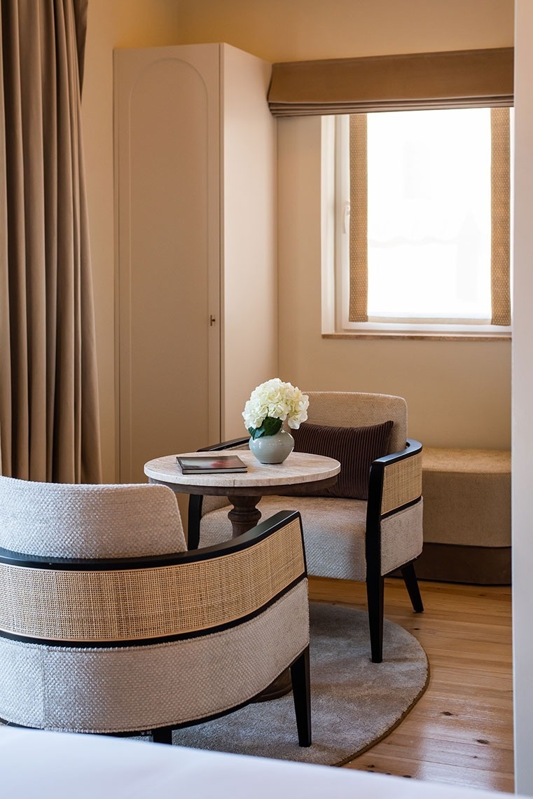 Área de estar aconchegante em quarto de hotel, com poltronas, mesa de centro e luz natural.