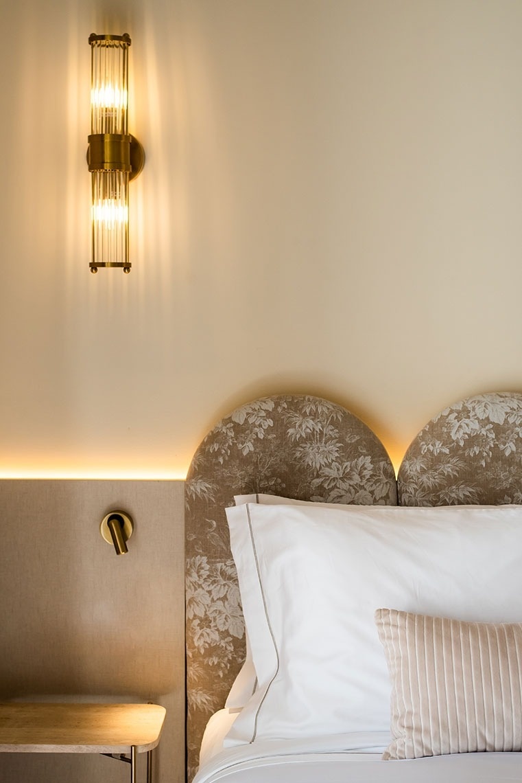 Cama de hotel elegante com cabeceira floral, luminária dourada e iluminação ambiente acolhedora.