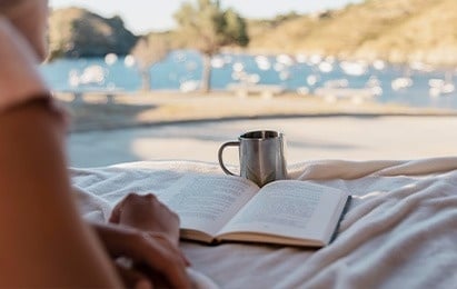 una mujer está sentada en una cama leyendo un libro junto a una taza de café .