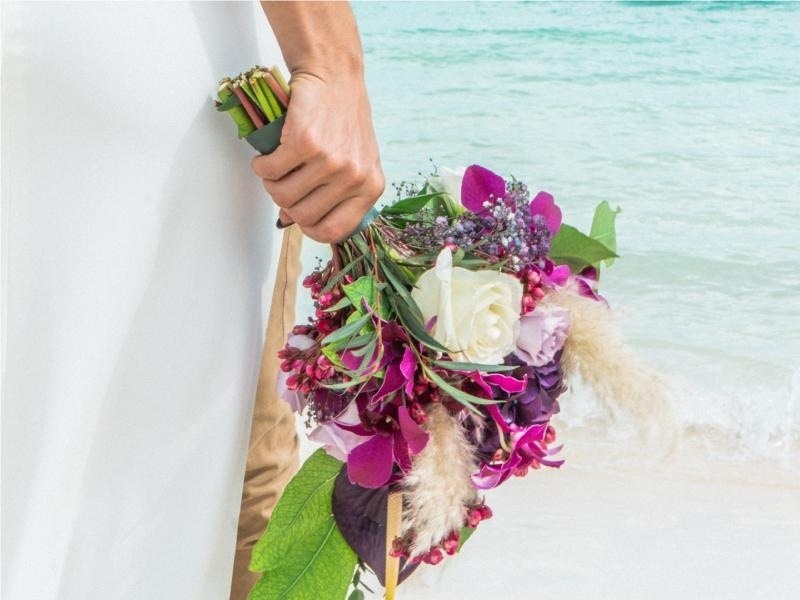 una novia sostiene un ramo de flores moradas y blancas en la playa