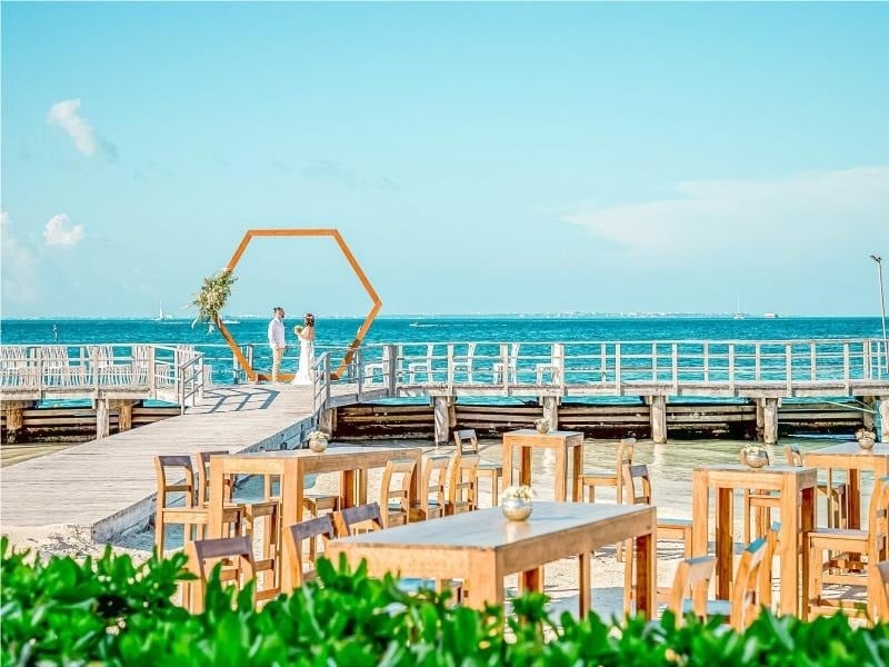 una novia y un novio se paran en un muelle con mesas y sillas frente al océano .