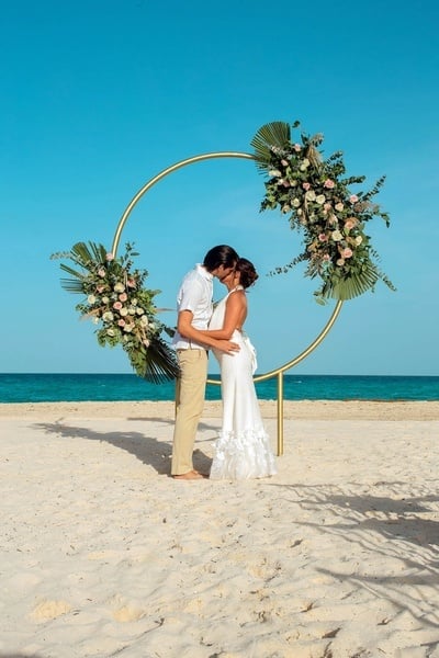 a bride and groom kiss under a floral arch on the beach