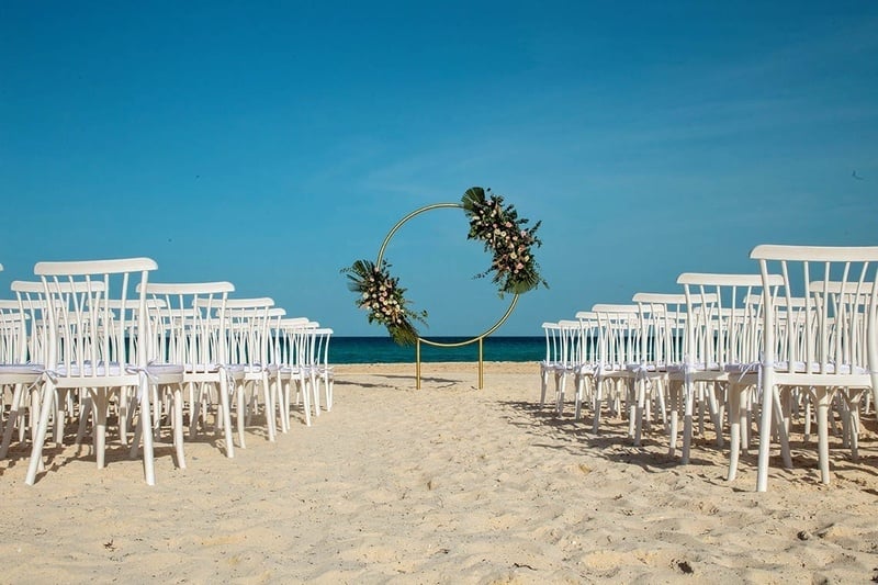 a row of white chairs on a beach with a circle of flowers in the background