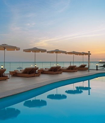 una piscina llena de sillas y sombrillas con vistas al océano al atardecer