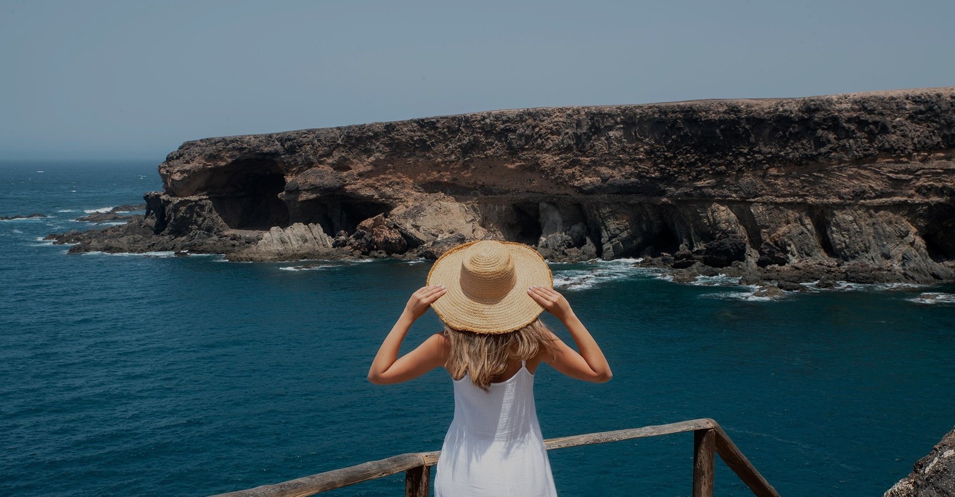 a woman in a white dress and straw hat looks out over the ocean