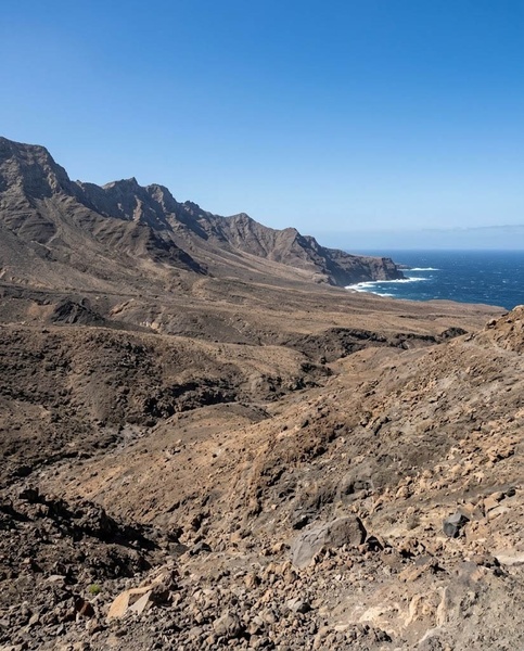 Un vaste paysage côtier aride révèle des montagnes rocheuses plongeant dans l'océan bleu sous un ciel dégagé.