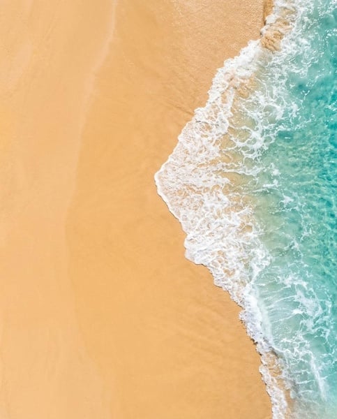 Une plage de sable fin est bordée par une mer turquoise aux vagues légères sous un ciel bleu parsemé de nuages.