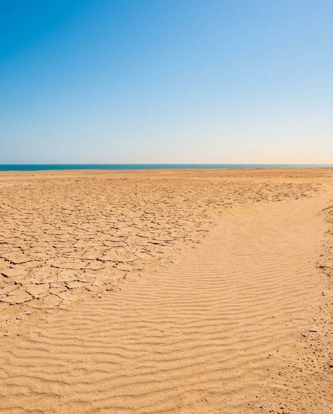 L'image présente un paysage aride de terre craquelée et de sable ondulé rejoignant une mer bleue sous un ciel clair.