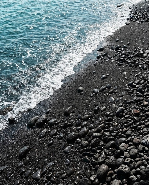 Une mer d'un bleu-vert éclatant s'écrase en vagues moussantes sur une plage de galets noirs.