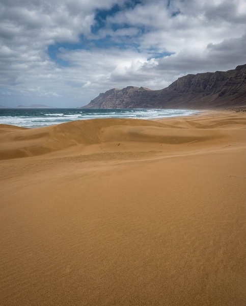 Vastes dunes de sable doré bordent un océan agité avec des montagnes en arrière-plan sous un ciel nuageux.