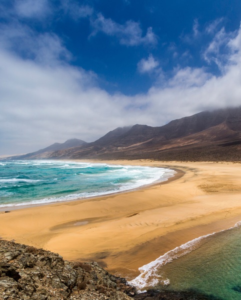 Vastes dunes de sable doré bordent un océan agité avec des montagnes en arrière-plan sous un ciel nuageux.