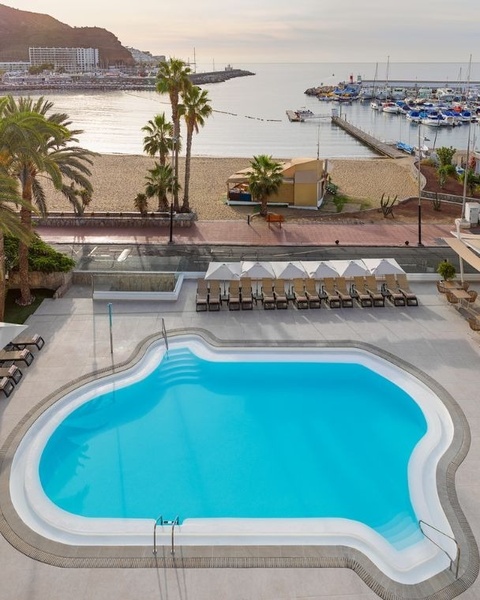 An aerial view captures a uniquely shaped swimming pool in the foreground, with a sandy beach, palm trees, and a bustling marina leading out to the ocean and distant hills under a soft, overcast sky.