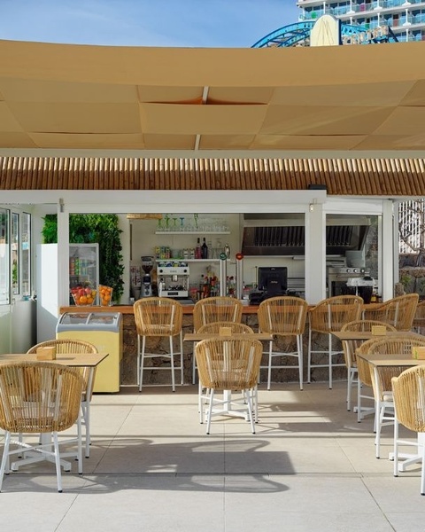 A bright, open-air cafe features a service counter with rattan bar stools, along with tables and chairs on a paved patio, all shaded by a large beige canopy, with a multi-story building visible in the background.
