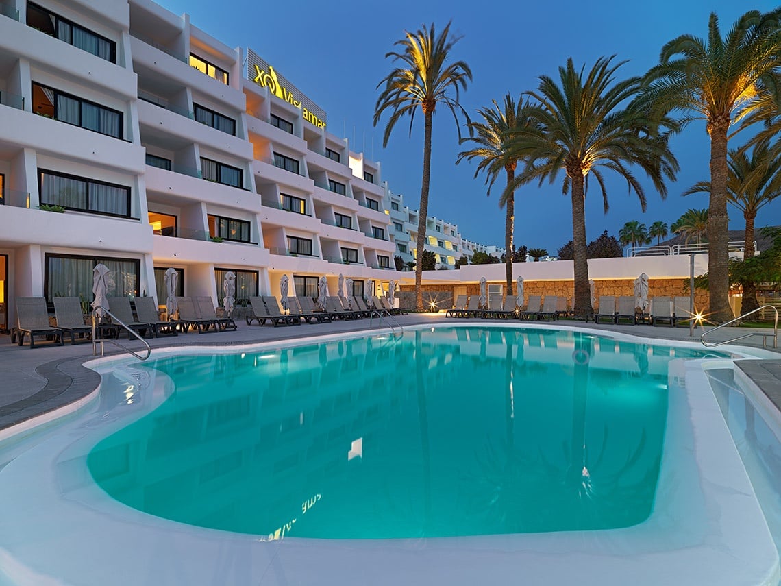 XQ Apartamentos Vistamar: piscina cristalina y palmeras bajo el atardecer.