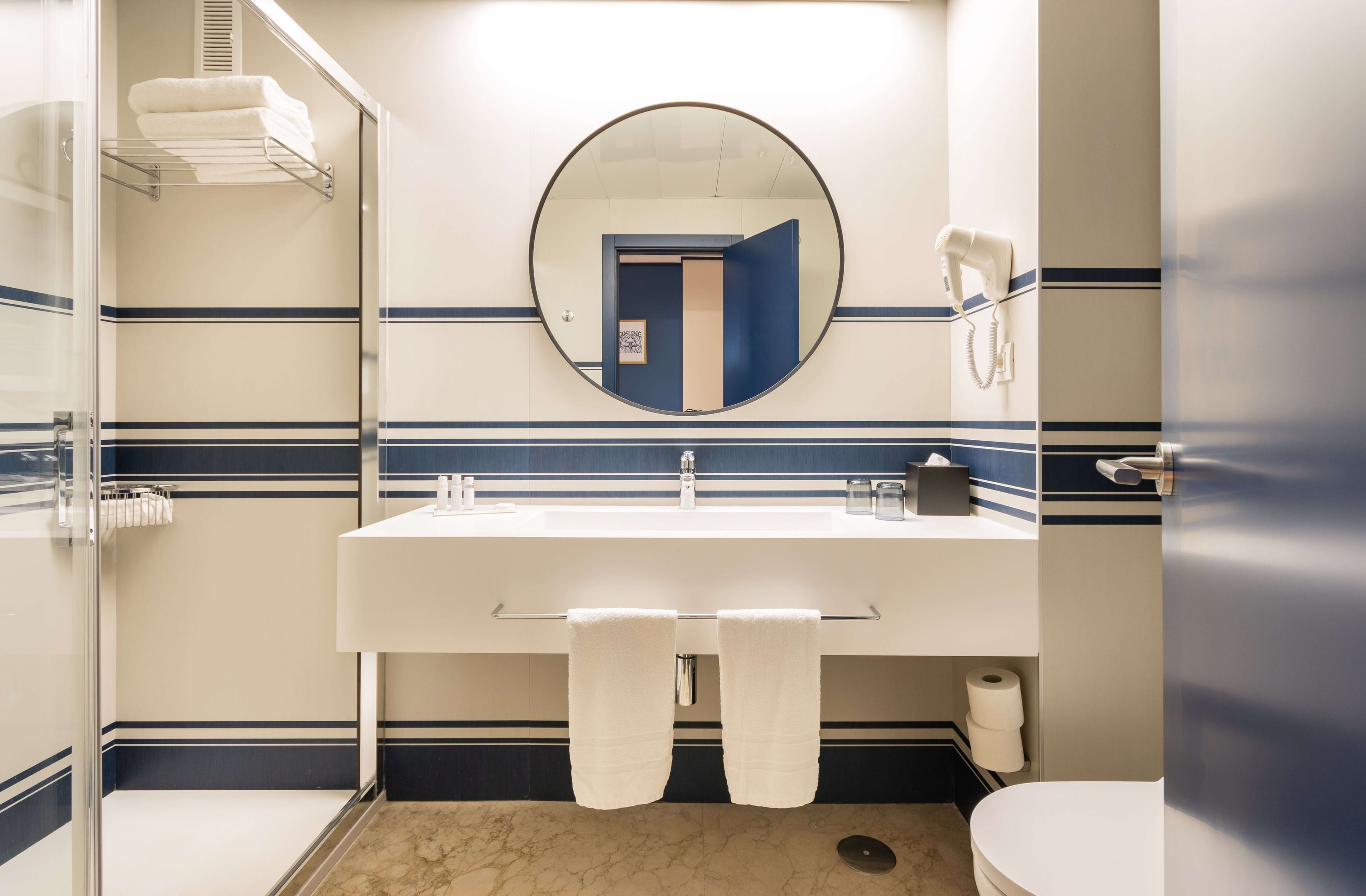 Un baño moderno y luminoso presenta paredes con rayas azules y blancas, un lavabo con un espejo redondo, una ducha de cristal y un inodoro.