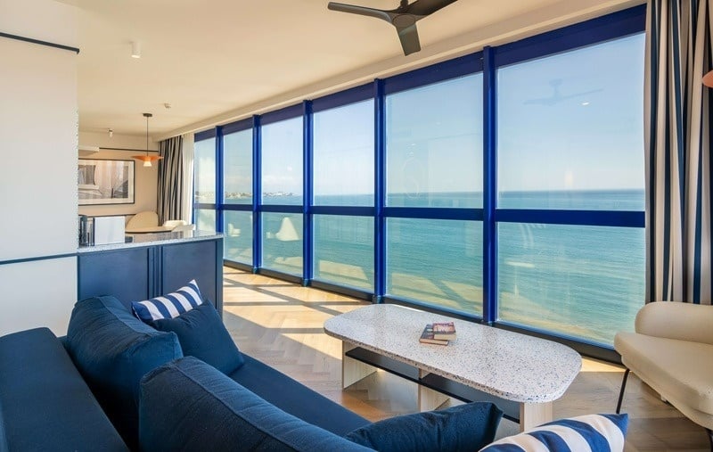 Un moderno salón con grandes ventanales y decoración en tonos azules, ofreciendo una impresionante vista al mar.