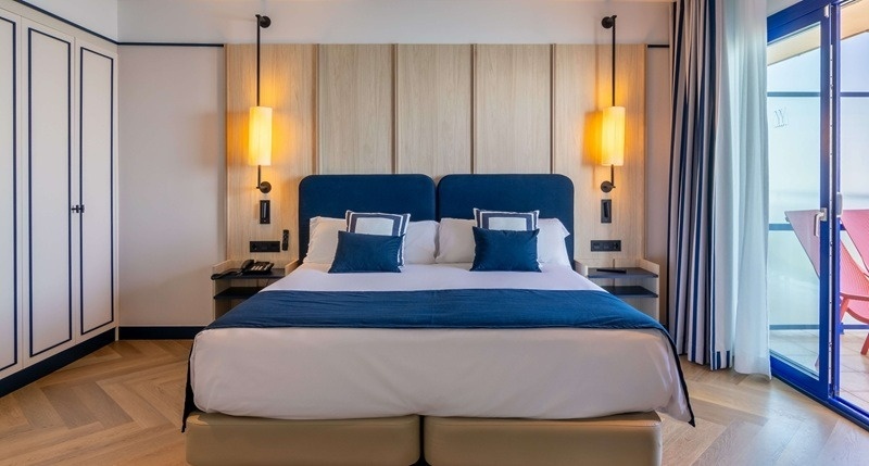 Una habitación de hotel moderna y luminosa con una cama grande de cabeceros azules, paneles de madera clara en la pared, un armario y acceso a un balcón con vistas.