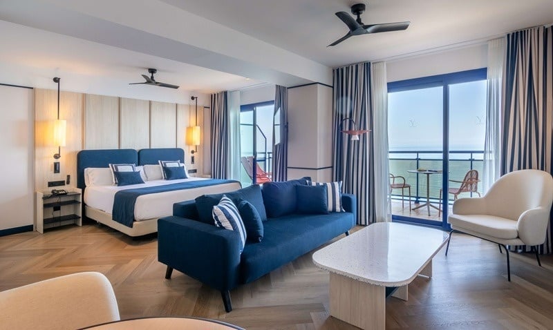 Moderna suite de hotel con diseño náutico en azul y blanco, que cuenta con cama, sofá y balcón con vistas al mar.