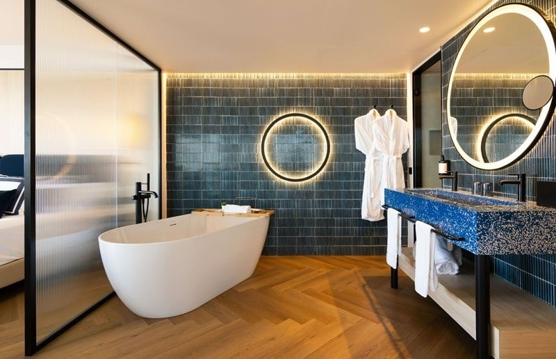 Este baño contemporáneo presenta una bañera ovalada, un llamativo lavabo doble azul, una pared de azulejos texturizados con iluminación ambiental y un suelo de madera en espiga, separado de la habitación por un panel de vidrio estriado.