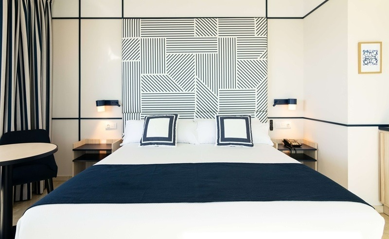 Una moderna habitación de hotel con una cama doble y un cabecero geométrico en blanco y negro, complementada con ropa de cama blanca y azul marino y cortinas a rayas.