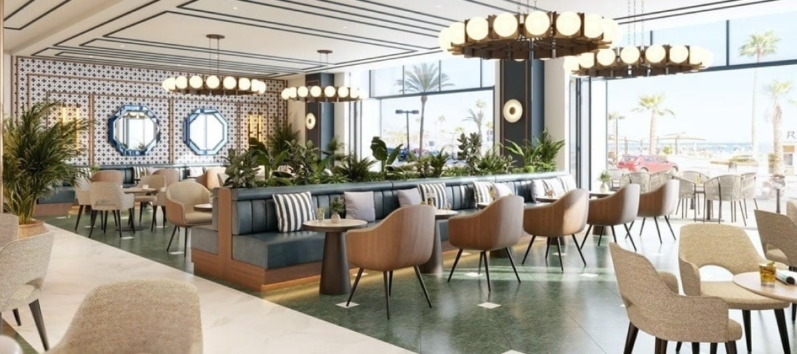 Un moderno y luminoso restaurante o cafetería con un diseño interior elegante, cómodos asientos, abundante vegetación y grandes ventanales con vistas a la playa y al mar.