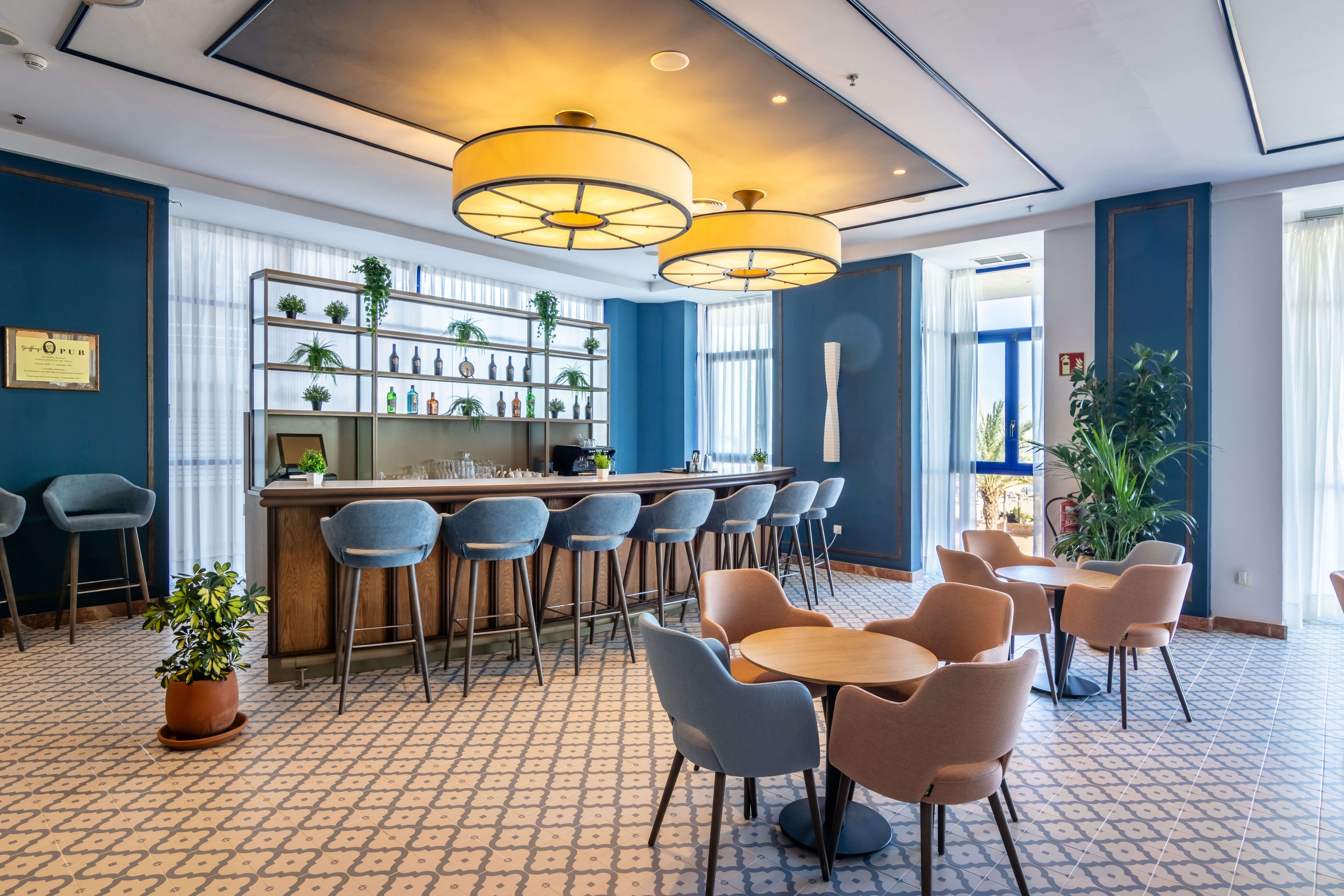 Un bar d'hôtel moderne est aménagé avec un long comptoir en bois, des tabourets bleus, un sol à motifs et plusieurs tables rondes avec des fauteuils, le tout sous de grands luminaires circulaires.