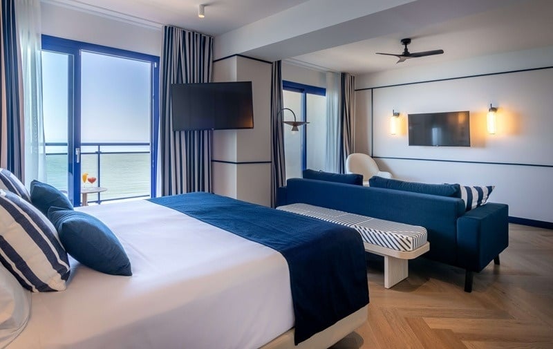 Una moderna habitación de hotel con decoración náutica en tonos azules y blancos, y un balcón con vistas al mar.