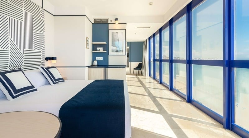 Una luminosa habitación de hotel moderna, decorada en tonos azules y blancos, cuenta con una cama grande y una pared de ventanales que ofrecen vistas panorámicas.