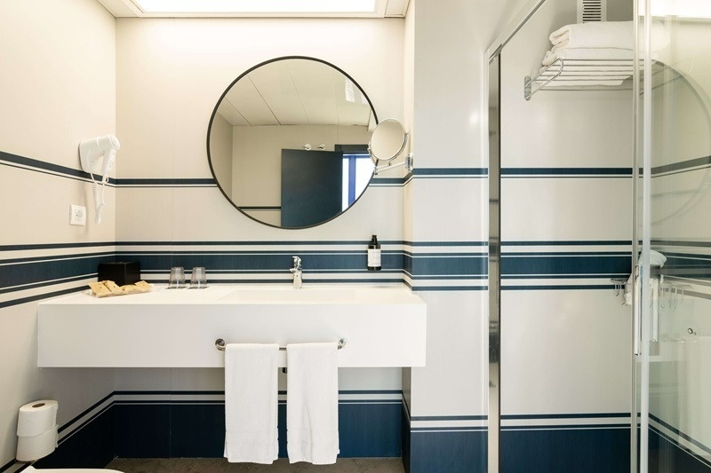 Un baño moderno con azulejos a rayas blancas y azul marino, equipado con un lavabo suspendido, un espejo circular y una cabina de ducha.