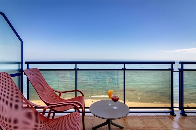Un balcón con dos sillas y bebidas ofrece una vista al mar y a la playa bajo un cielo azul.