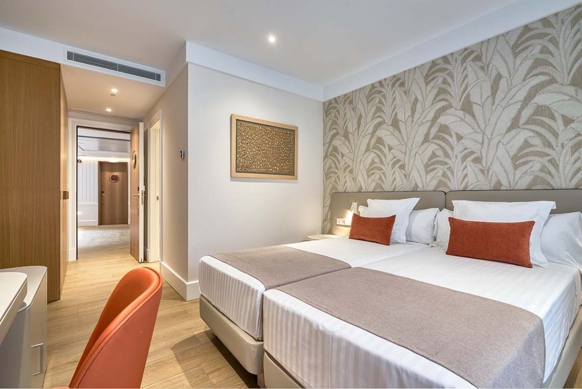 Una habitación de hotel luminosa con dos camas individuales, papel tapiz de hojas detrás de la cabecera y un pasillo que se adentra hacia el fondo.