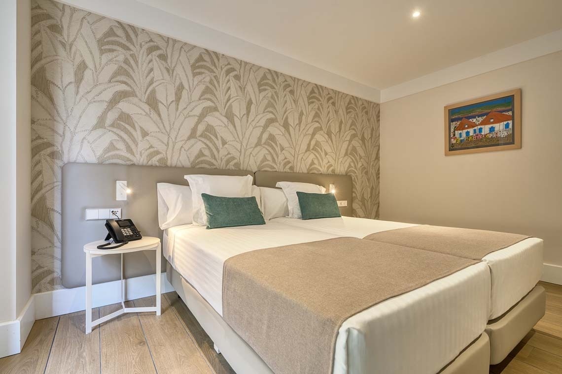 Se muestra una habitación de hotel con dos camas individuales, un cabecero acolchado, papel tapiz con diseño de hojas y un cuadro colorido en la pared.