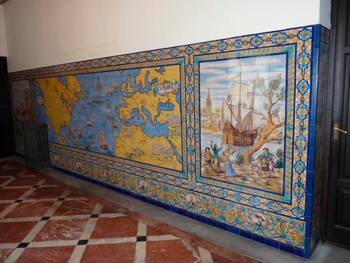 Una pared está adornada con coloridos azulejos de cerámica que representan un mapa antiguo del Atlántico con barcos y una vibrante escena portuaria, sobre un suelo de mármol.