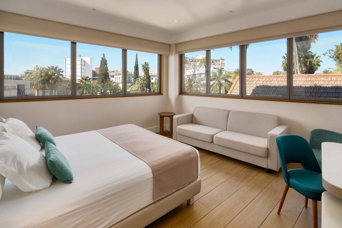 Una habitación moderna y luminosa con una cama, un sofá y amplios ventanales que ofrecen vistas a un paisaje urbano de palmeras y edificios.
