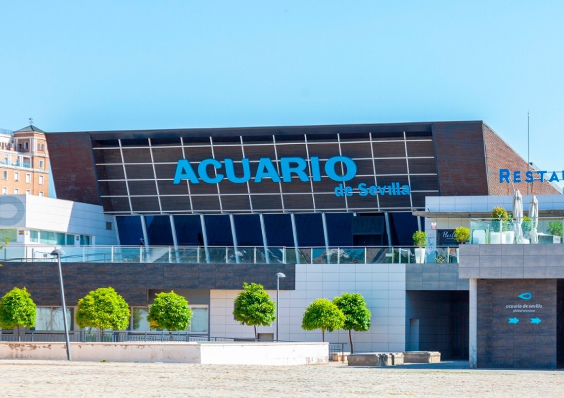 Fachada del Acuario de Sevilla con su nombre en letras azules brillantes y parte del letrero de un restaurante, bajo un cielo azul claro.