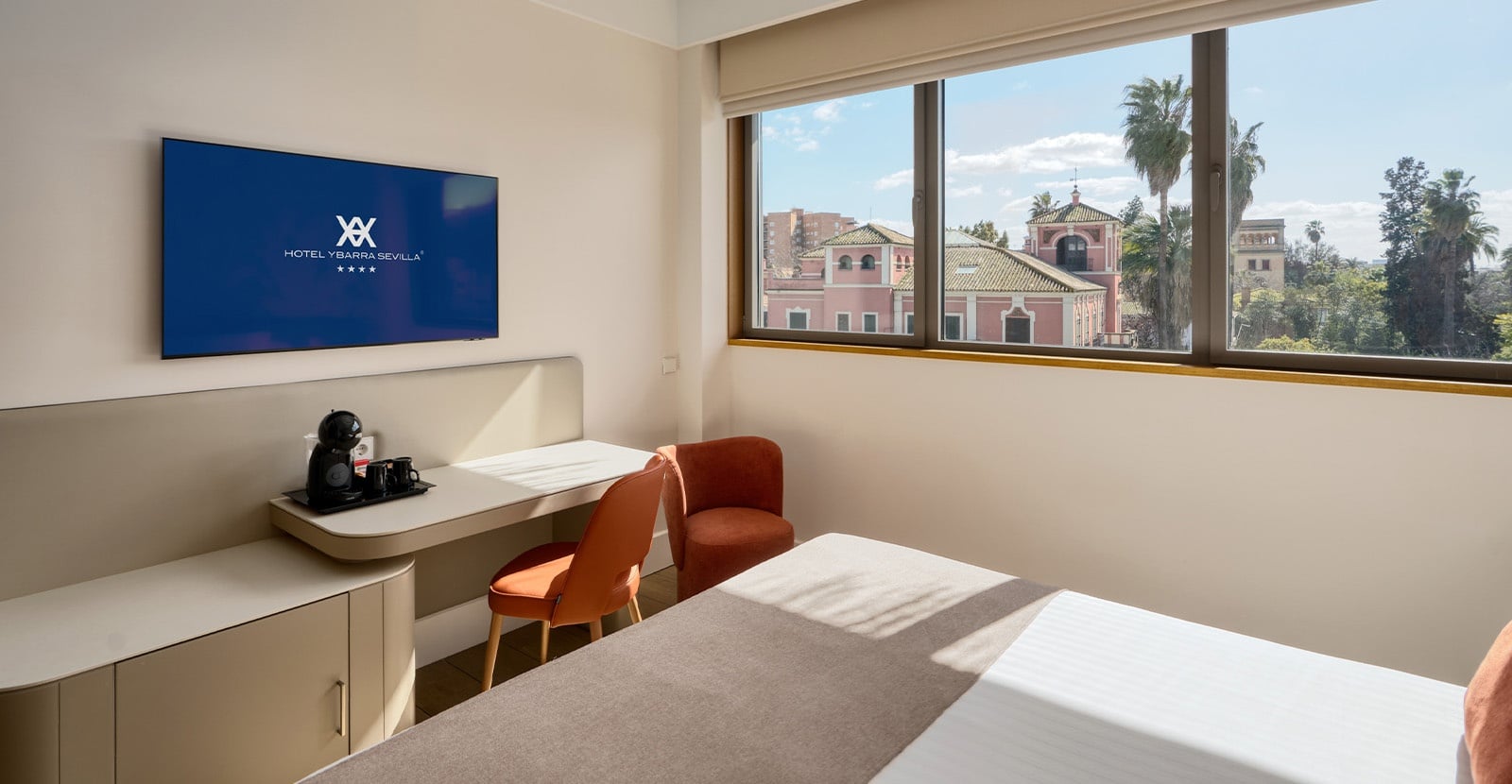 Una moderna habitación de hotel con una cama, un escritorio con dos sillas, una cafetera, un televisor y una ventana con vistas a edificios y palmeras.