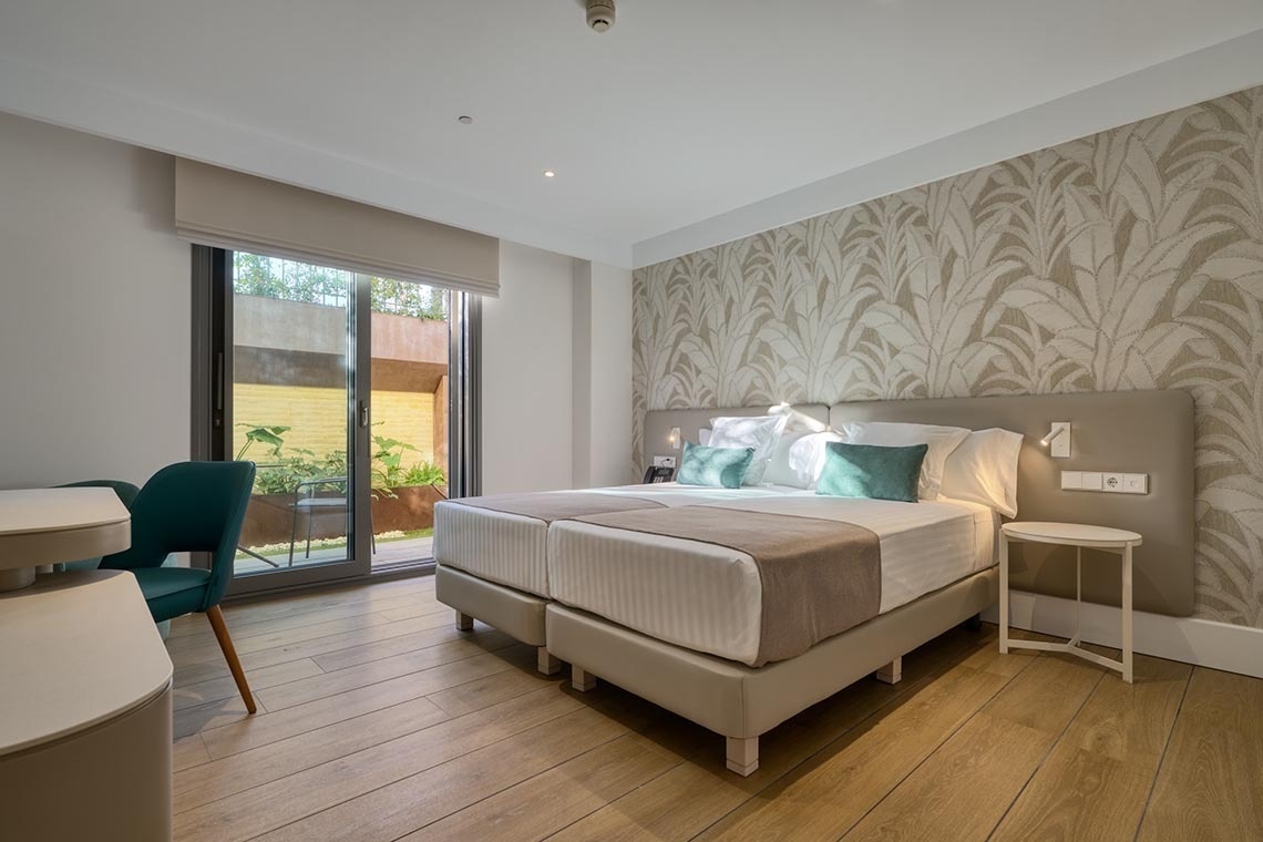 Una moderna habitación de hotel presenta dos camas individuales con cabecero tapizado y cojines de color verde azulado, suelo de madera, una pared de acento con papel pintado de hojas y una puerta de cristal que da a un pequeño patio exterior con vegetación.