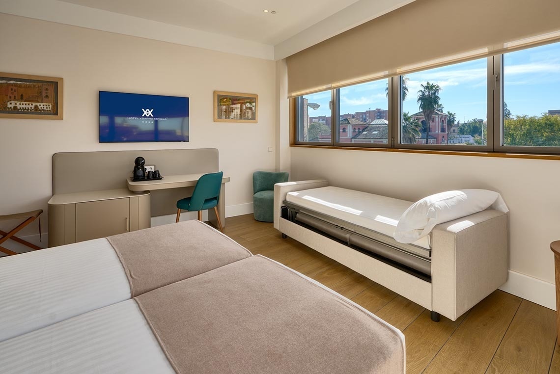 Habitación de hotel moderna y luminosa con dos camas, sofá cama, televisor de pantalla plana y amplias ventanas con vista al exterior.