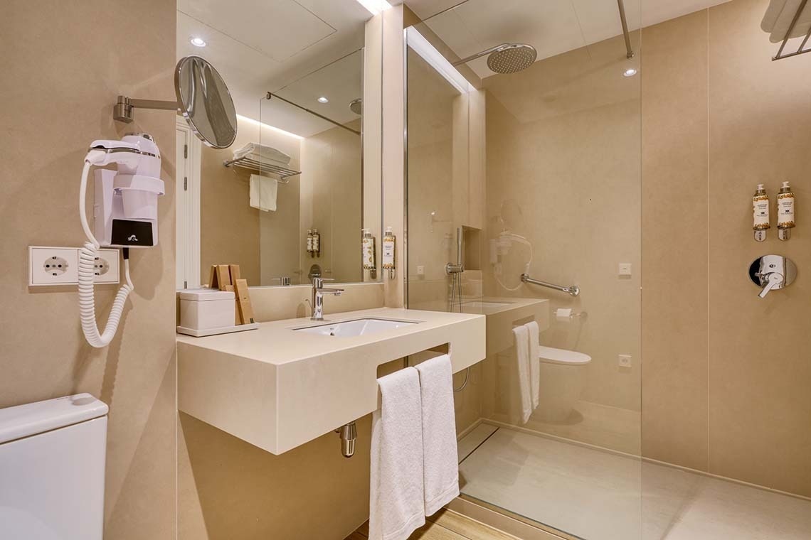 Un baño moderno y funcional en tonos neutros, equipado con lavabo, secador de pelo montado en la pared y una ducha a ras de suelo con mampara de cristal.