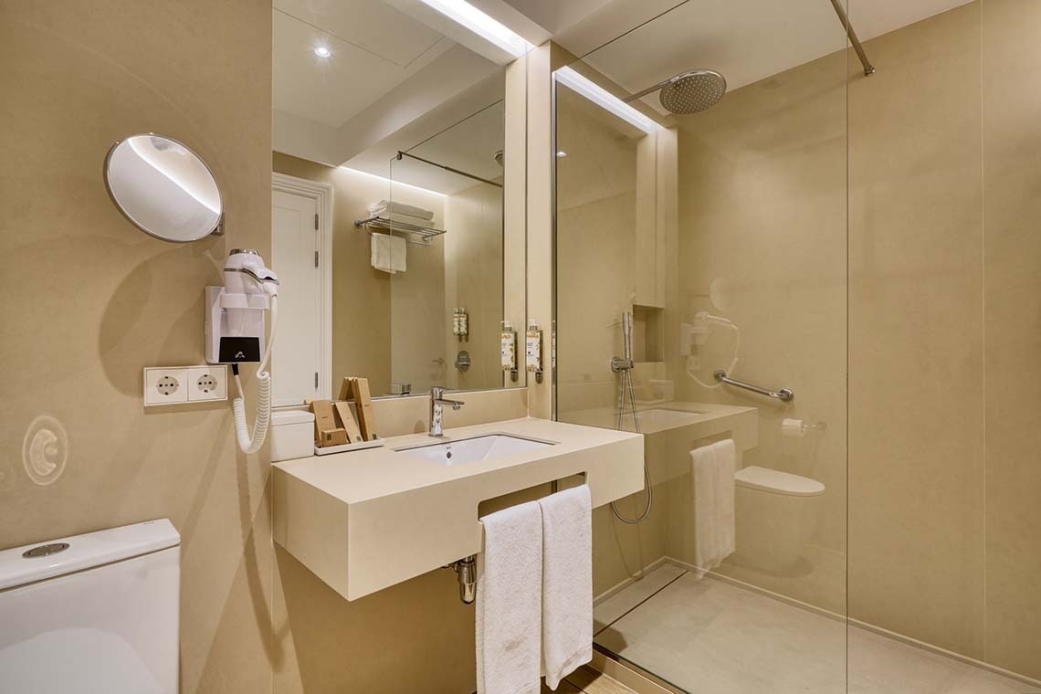 Un moderno baño de tonos beige con un lavabo flotante, un espejo grande, un inodoro y una ducha a ras de suelo con mampara de cristal.