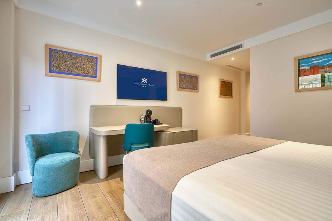 Una moderna habitación de hotel con una cama, escritorio, televisión montada en la pared y obras de arte decorando las paredes.