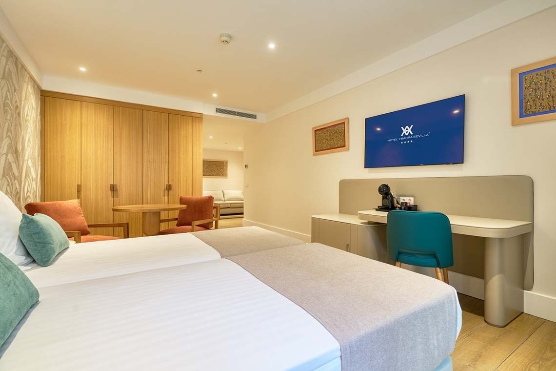 Una moderna habitación de hotel presenta dos camas individuales, un escritorio con televisor, un armario de madera y una zona de comedor con sillas naranjas.