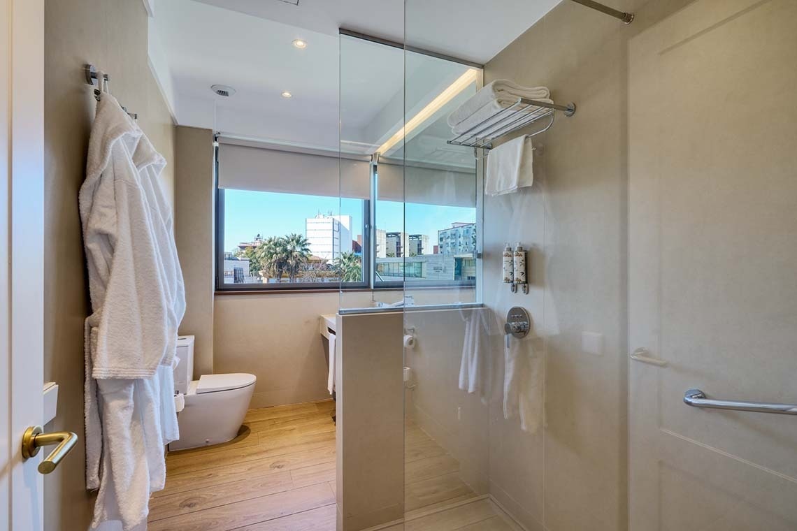 Este es un baño moderno con una ducha de cristal, un inodoro, y una ventana con vista a la ciudad, con toallas y albornoces colgados.