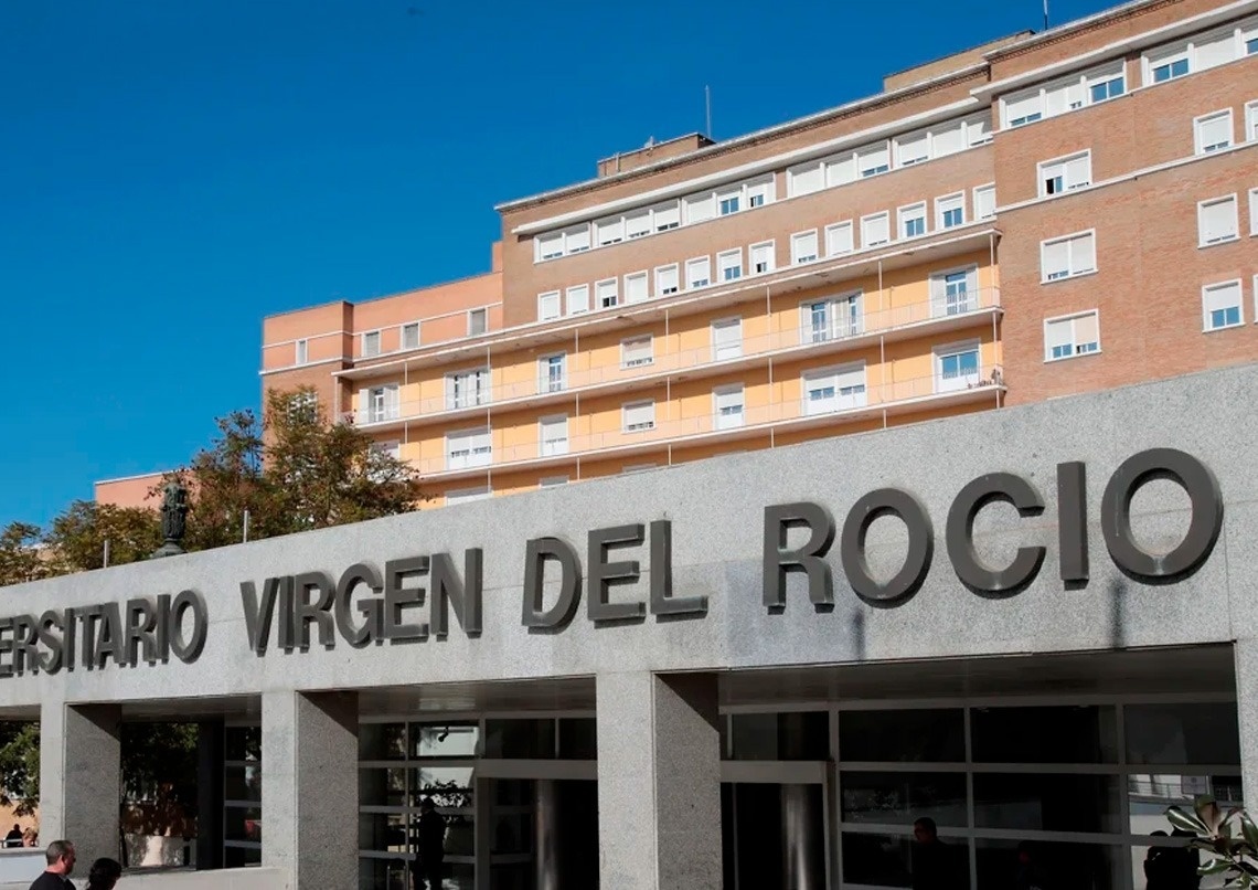 La imagen muestra la fachada del Hospital Universitario Virgen del Rocío, con su nombre destacado en letras grandes sobre la entrada principal, bajo un cielo azul claro.