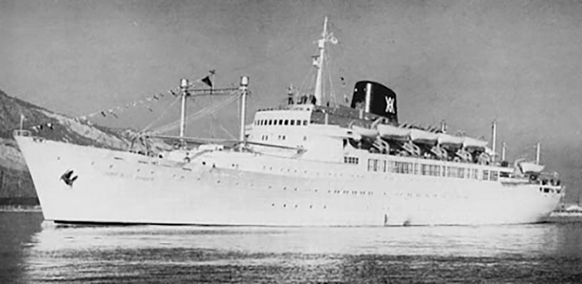 Una fotografía en blanco y negro muestra un transatlántico blanco con una chimenea oscura y banderines, navegando en aguas tranquilas frente a montañas.
