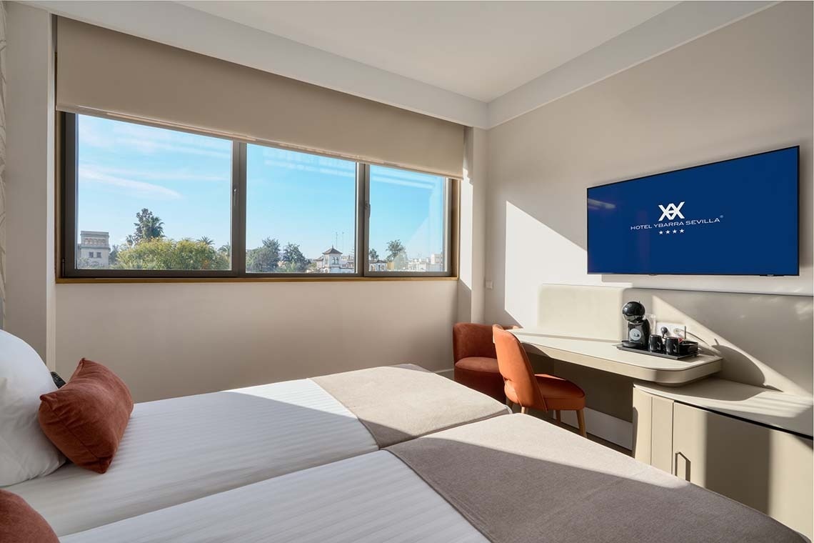 Una habitación de hotel moderna y luminosa con dos camas individuales, una gran ventana con vista a la ciudad y una zona de escritorio con televisión y cafetera.