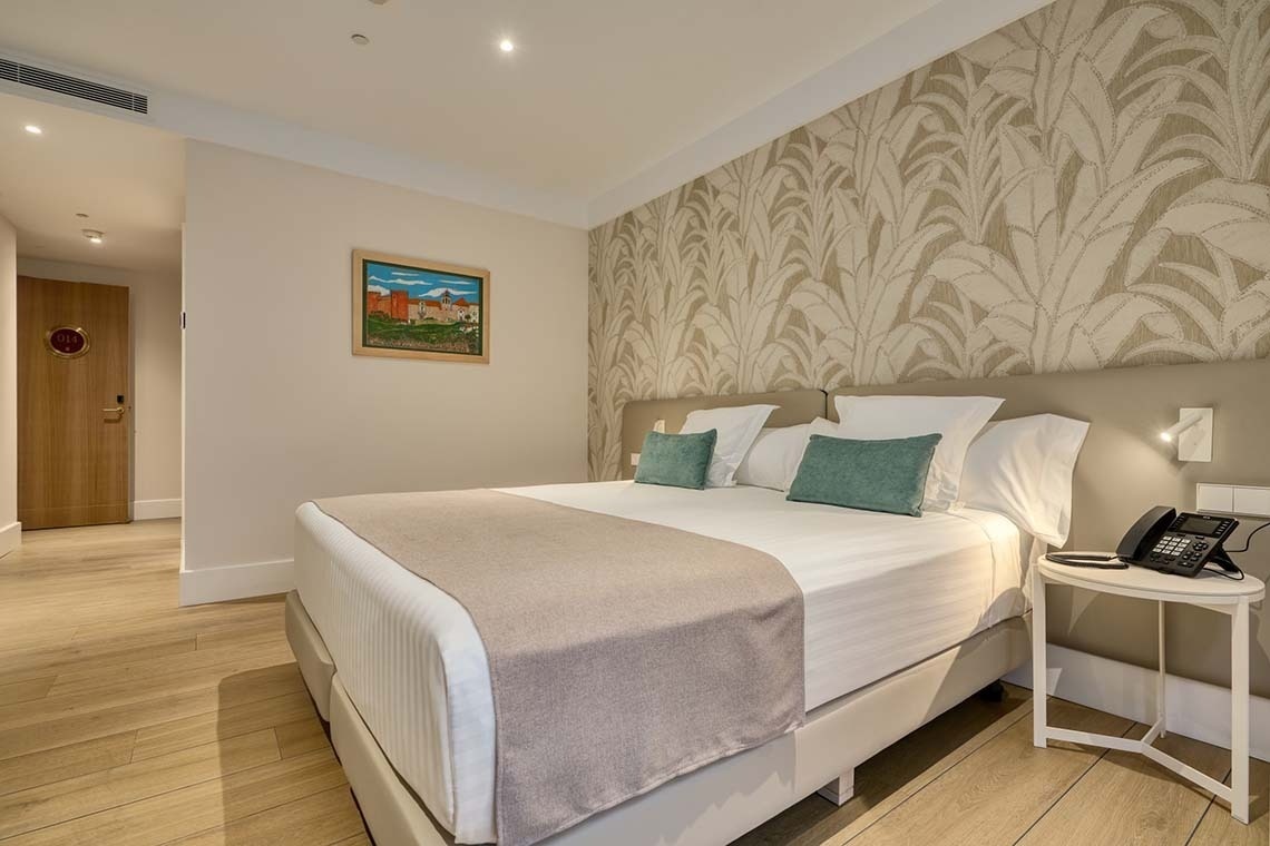 Una habitación de hotel moderna y luminosa con una cama doble, cojines decorativos turquesa, papel tapiz con patrón de hojas y suelo de madera clara.